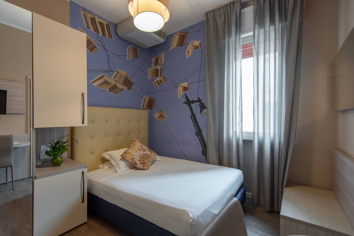 A modern hotel room with a bed, a small desk, a plant, and a unique wall decoration.