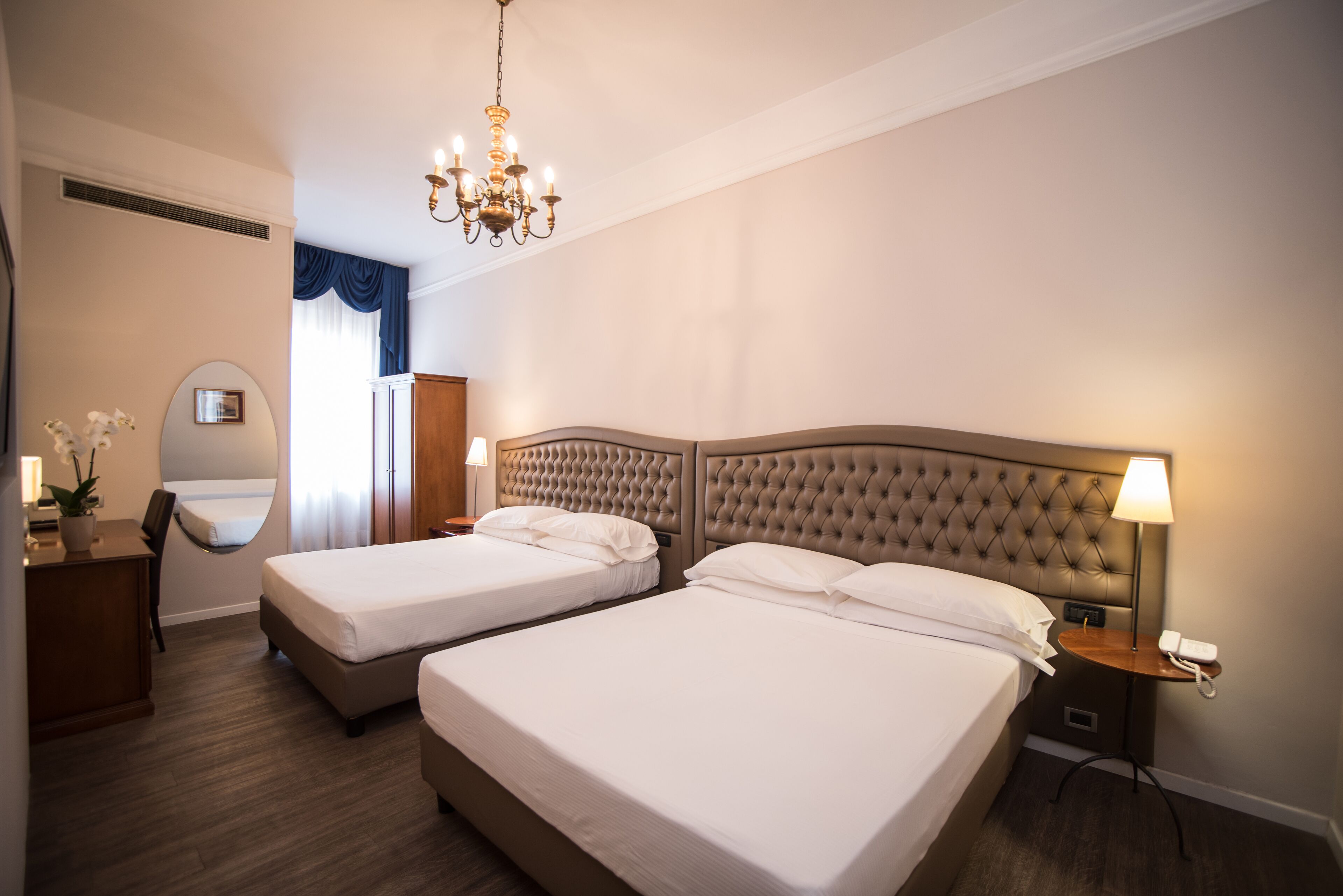 A hotel room with two beds, a desk, a mirror, a chandelier, and a bedside table with a lamp and phone.