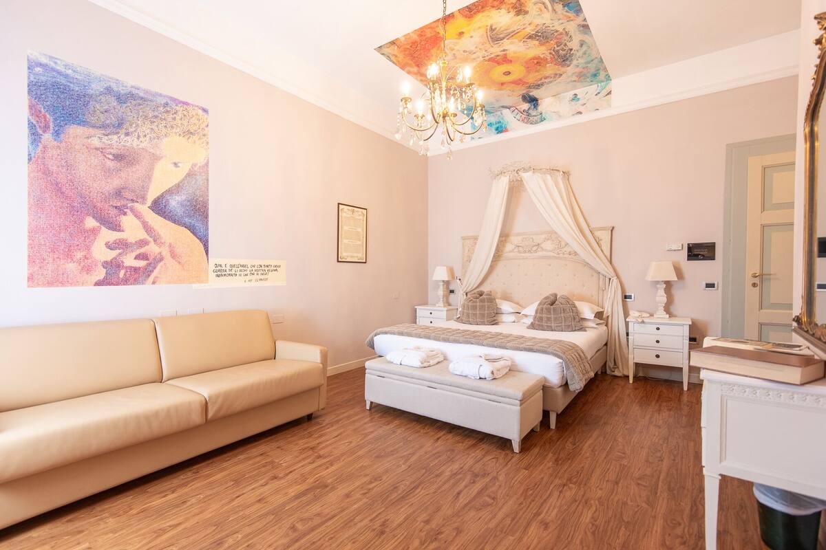 A bedroom with a four-poster bed, a bedside table, a nightstand, a bench, a large painting, and a chandelier.