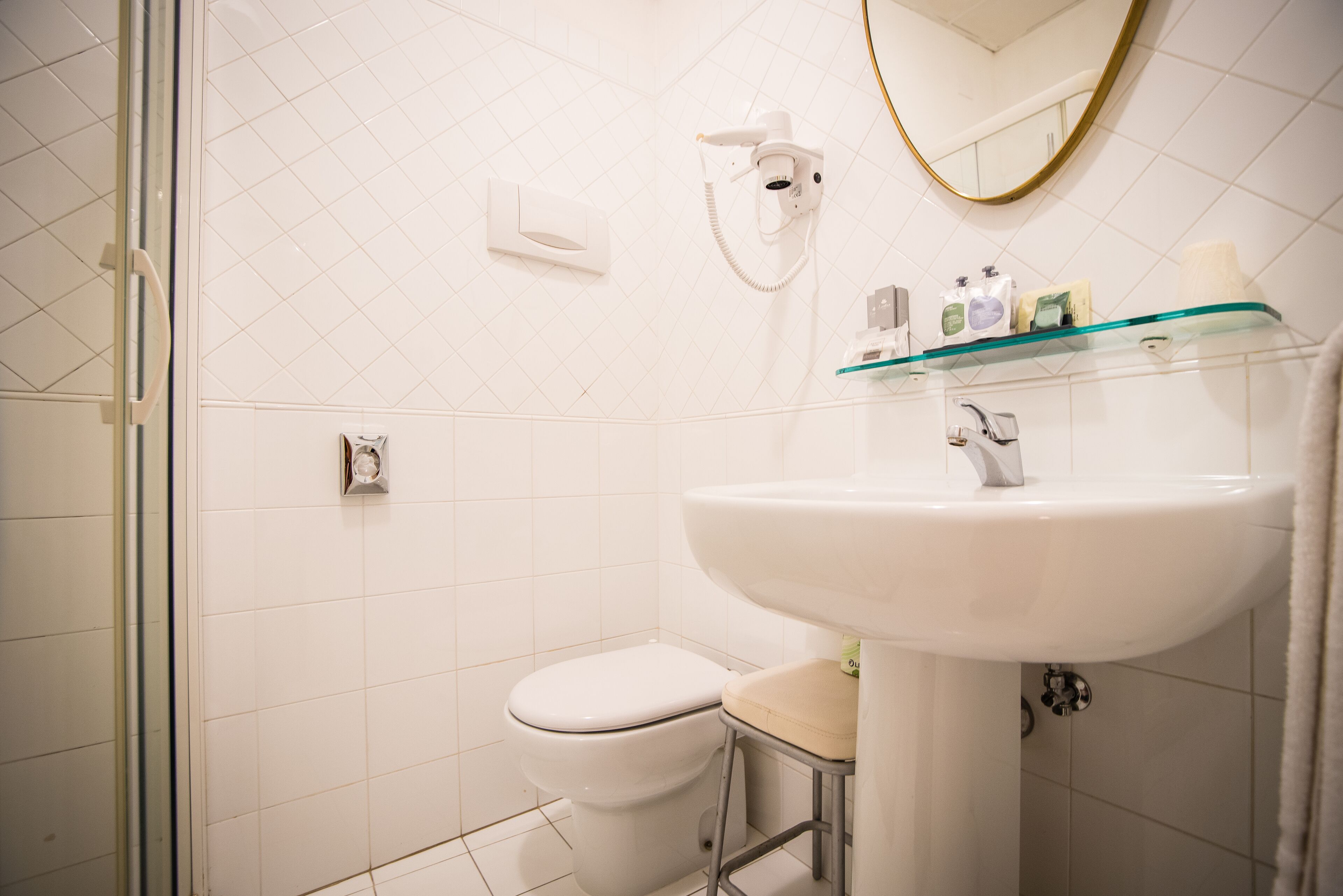 Un bagno con un water bianco, uno scaffale in vetro con articoli da toilette, un lavandino bianco e uno specchio con cornice dorata.