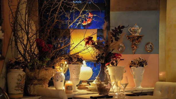 Una mesa de comedor con mantel blanco, rodeada de varios jarrones y velas, y una pared decorativa al fondo.