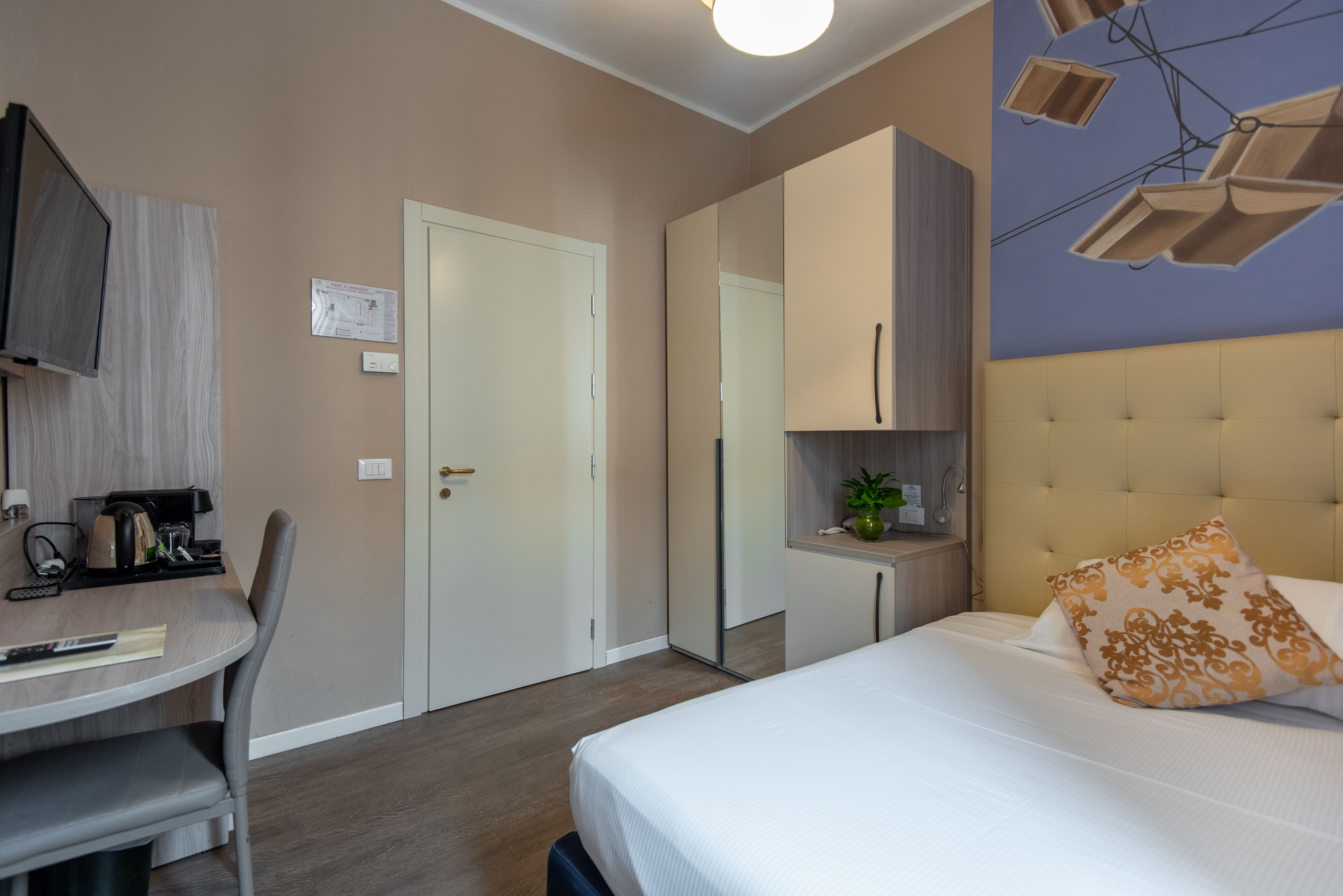 A hotel room with a bed, a desk with a chair, a wardrobe, and a mirror.