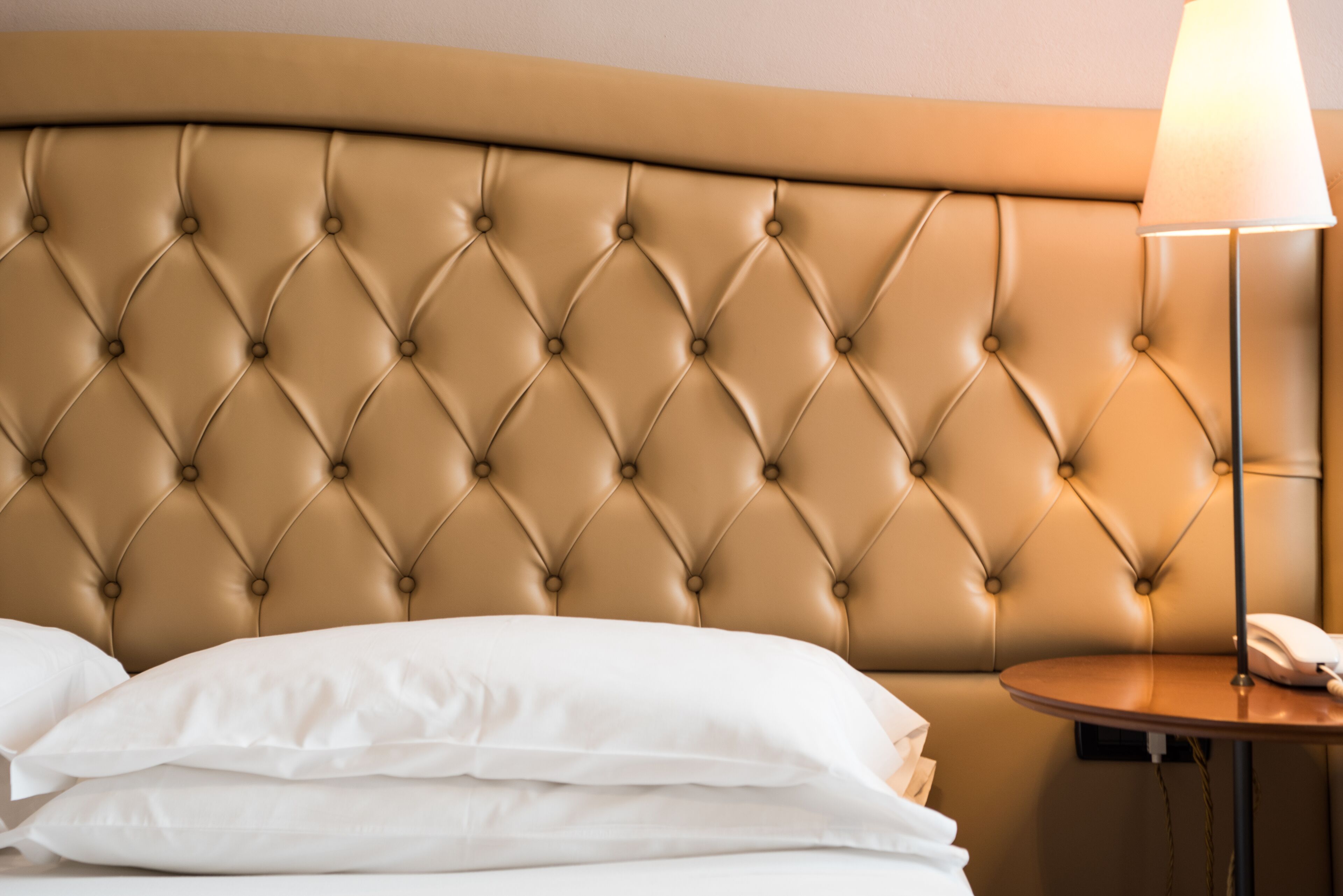 A hotel room with a tufted headboard, a round table with a lamp, and neatly made white pillows.