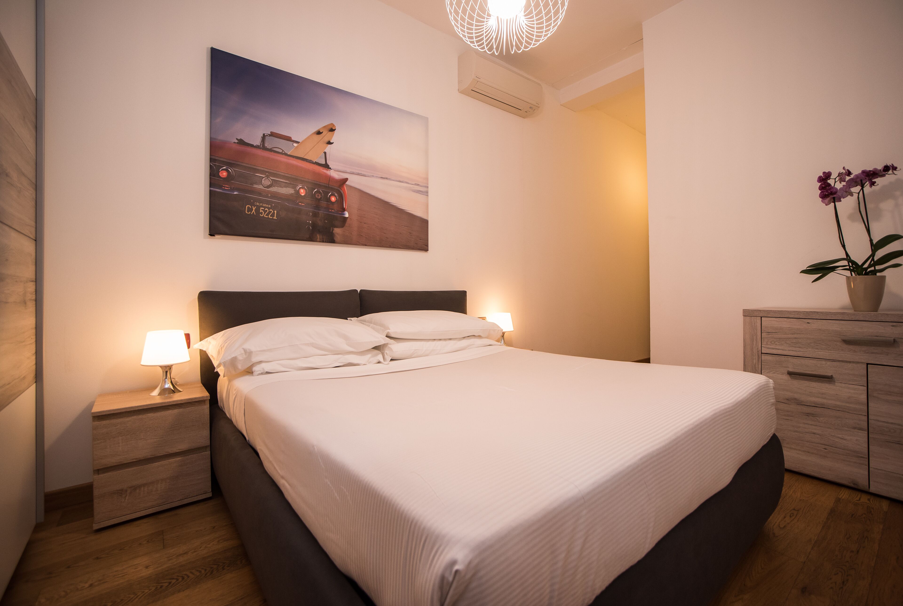 Una cama bien tendida con cabecera, dos mesitas de noche con lámparas, piso de madera, aire acondicionado de pared y una orquídea en maceta.