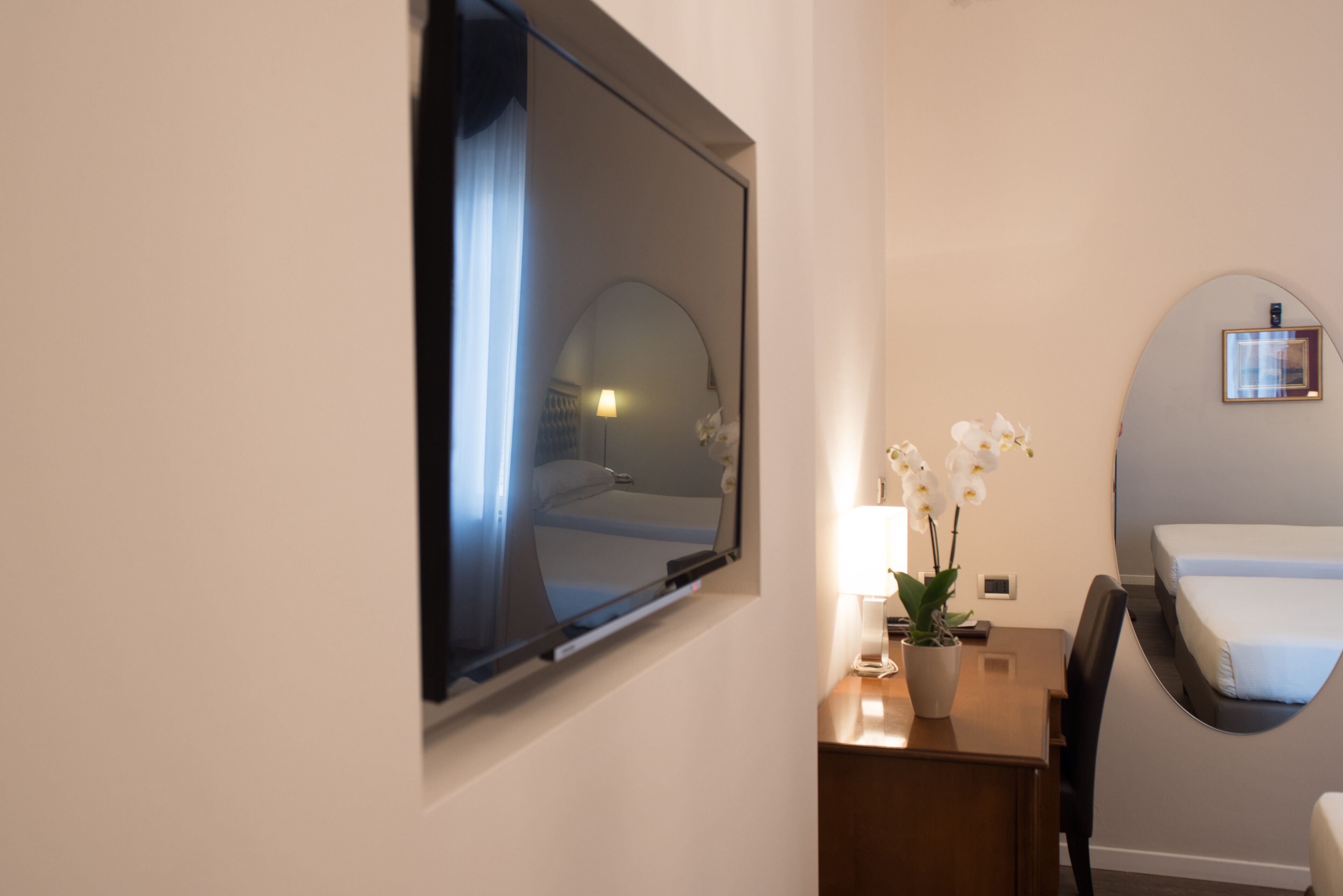 A hotel room with a bed, a desk with a lamp, a potted orchid, and a large mirror on the wall.