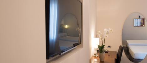 A hotel room with a bed, a desk with a lamp, a potted orchid, and a large mirror on the wall.