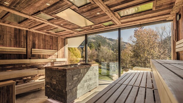 Una cabaña de madera con chimenea de piedra, bancos de madera y un ventanal con vistas a un paisaje montañoso.