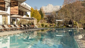 Piscina con sillas de descanso y sombrillas, rodeada por un edificio con detalles de madera y un paisaje montañoso de fondo.