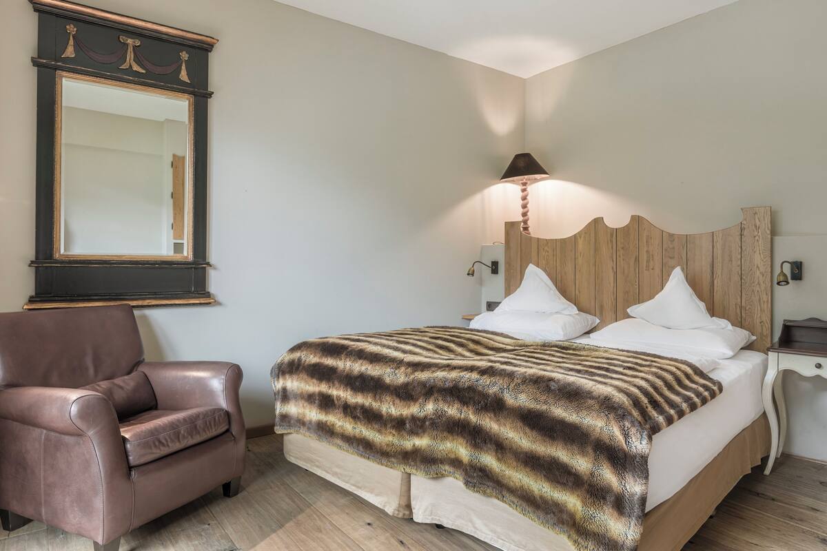 A bedroom with a bed, a leather armchair, a wooden headboard, and a mirror on the wall.
