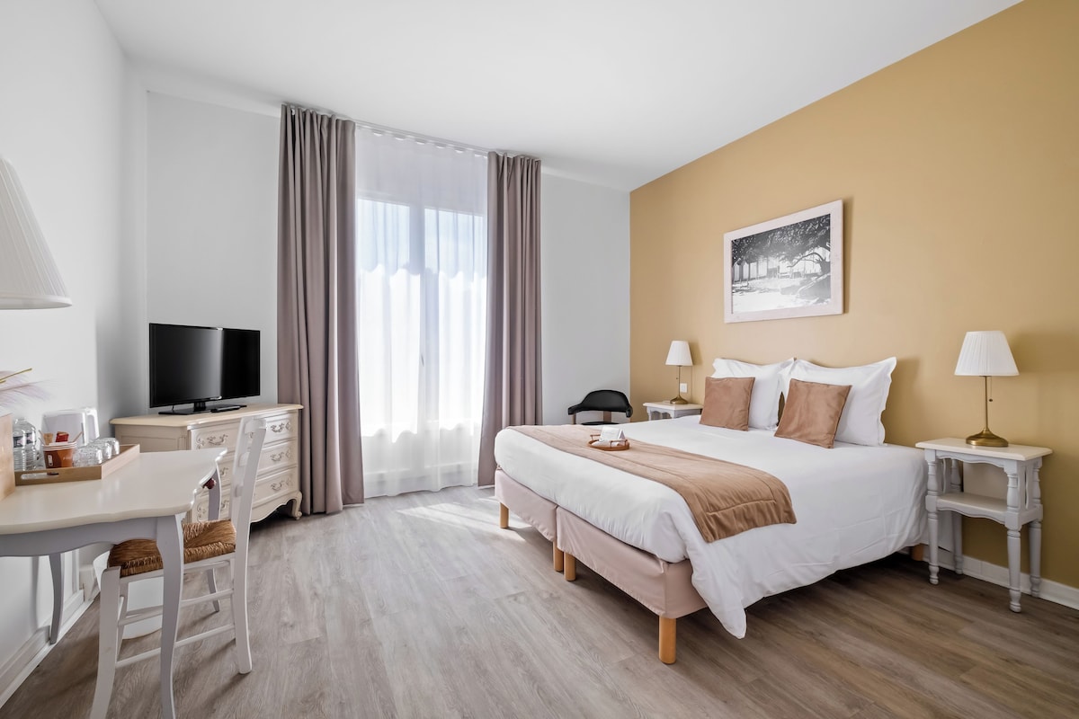 A hotel room with a bed, bedside tables, a desk with a television, and a chair.