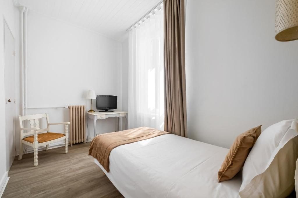 Une chambre d’hôtel avec un lit, une chaise, un bureau avec une télévision et une fenêtre avec des rideaux.