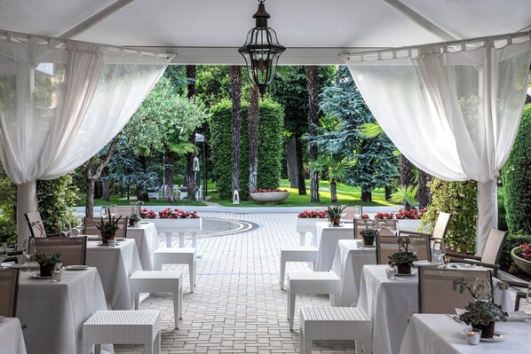 An outdoor dining area with white tables and chairs, surrounded by lush greenery and a paved walkway.