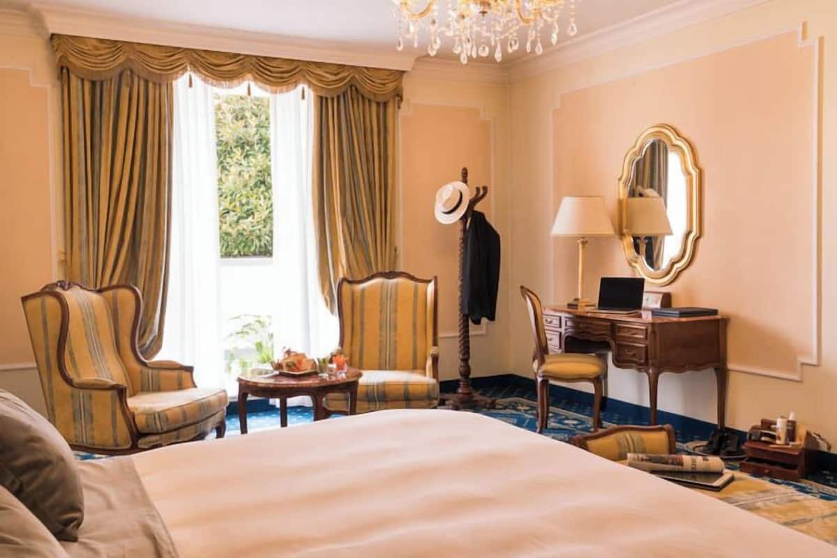 Ein Hotelzimmer mit einem großen Bett, zwei Sesseln, einem Schreibtisch mit Lampe, einem Spiegel und einem Fenster mit Vorhängen.