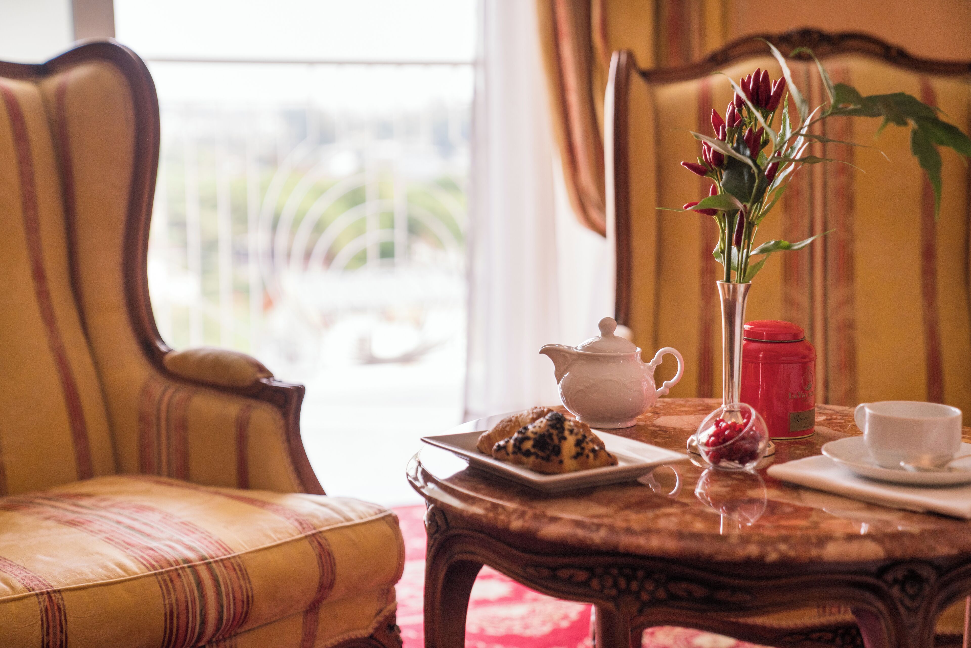 A dining table with a teapot, a plate of pastries, a vase with flowers, and a cup, set in a room with striped chairs and a view through a window.