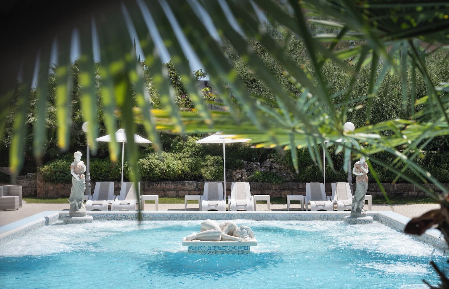 Une piscine entourée de verdure et de statues, avec des chaises longues et des parasols.