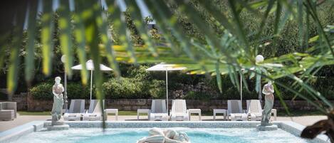Une piscine entourée de verdure et de statues, avec des chaises longues et des parasols.