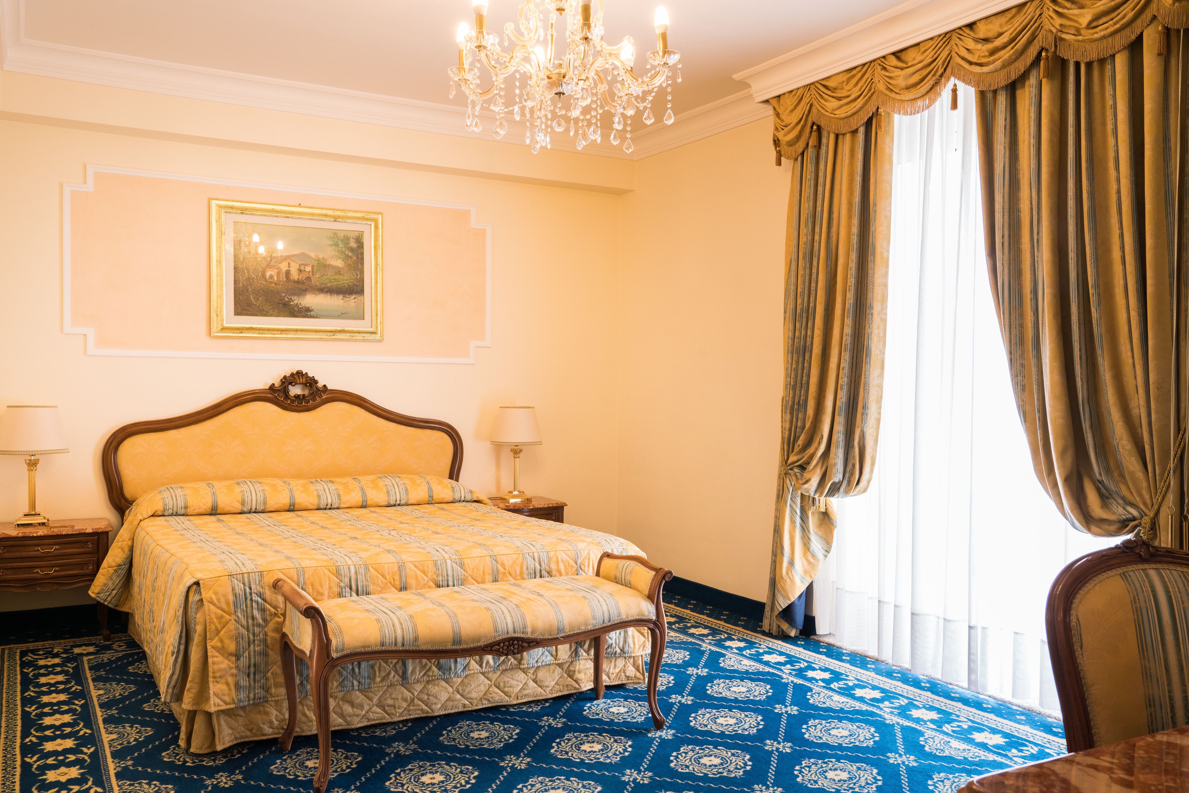 Quarto de hotel clássico com uma cama grande, um candelabro, um quadro na parede e vista através das cortinas.