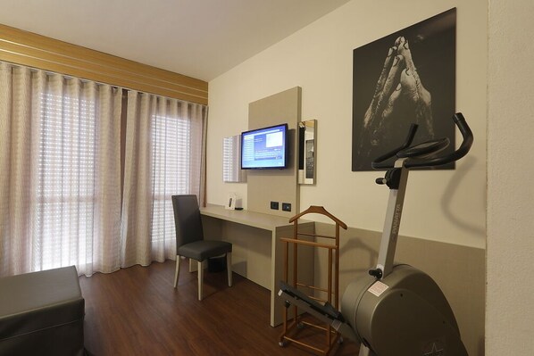 A hotel room with a wooden desk, a chair, a television, a treadmill, and a painting on the wall. - Hotel Campus (Collecchio)