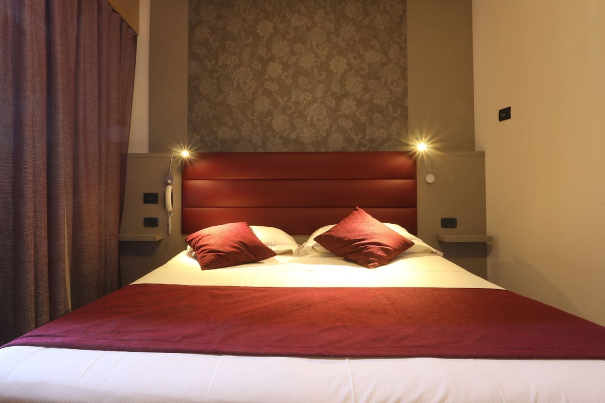 Una cama bien hecha con cabecera roja y almohadas a juego, dos lámparas de noche y un papel tapiz con diseño en la pared.