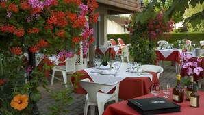 An outdoor dining area with tables set for two, surrounded by lush greenery and flowering plants. - LOGIS Chez Bach (Chaussin)