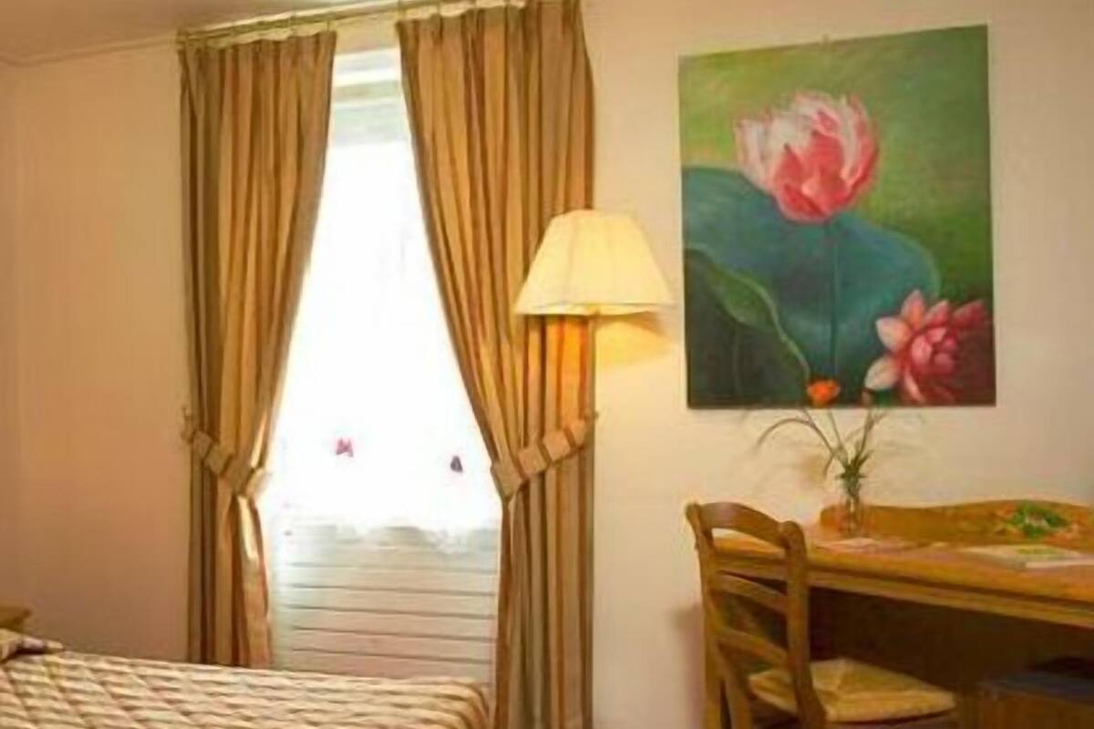 A hotel room with a bed, a desk, a chair, a lamp, and a painting of flowers on the wall.