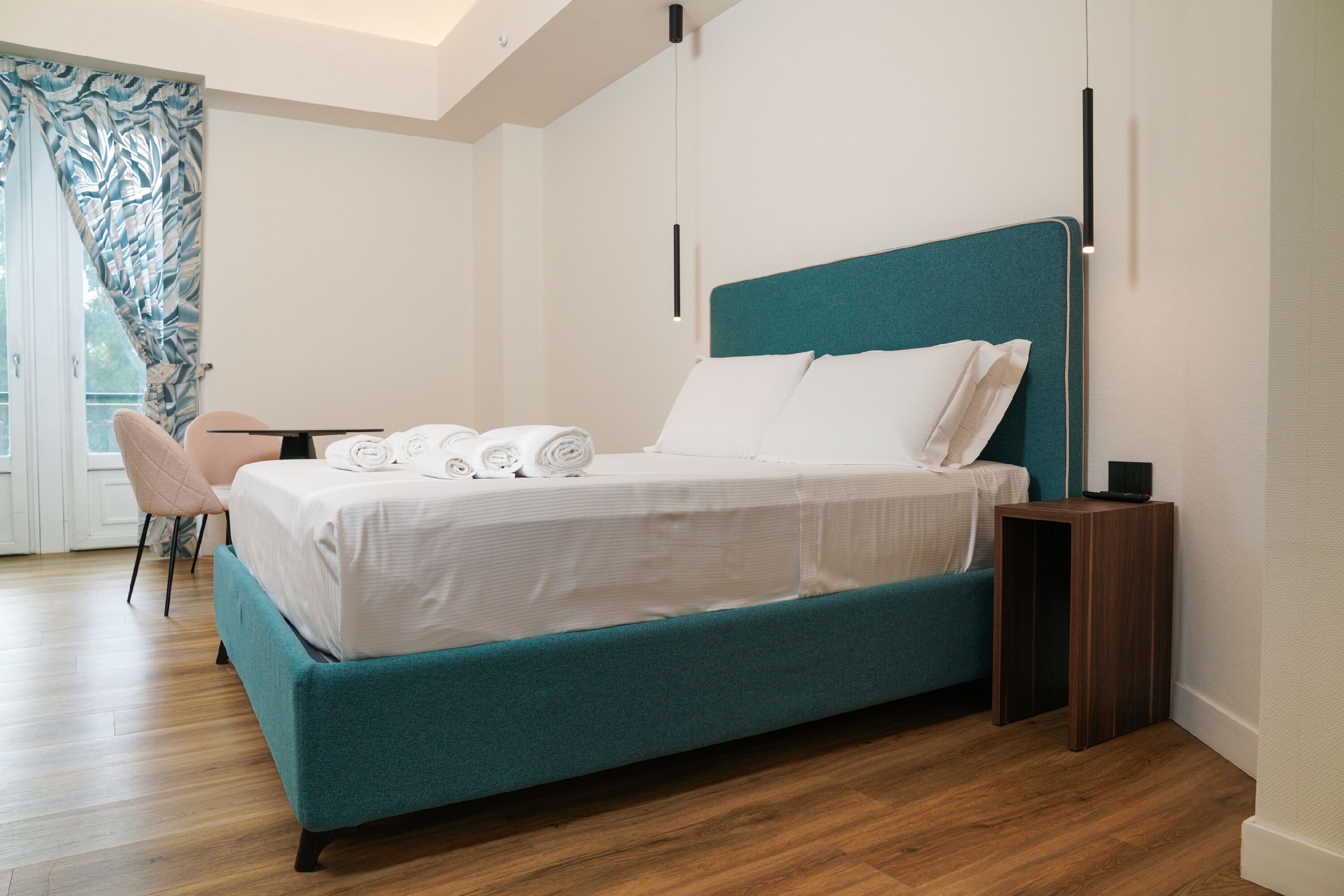 Un dormitorio moderno con una cama color turquesa, ropa de cama blanca, una mesita de noche de madera y una silla.