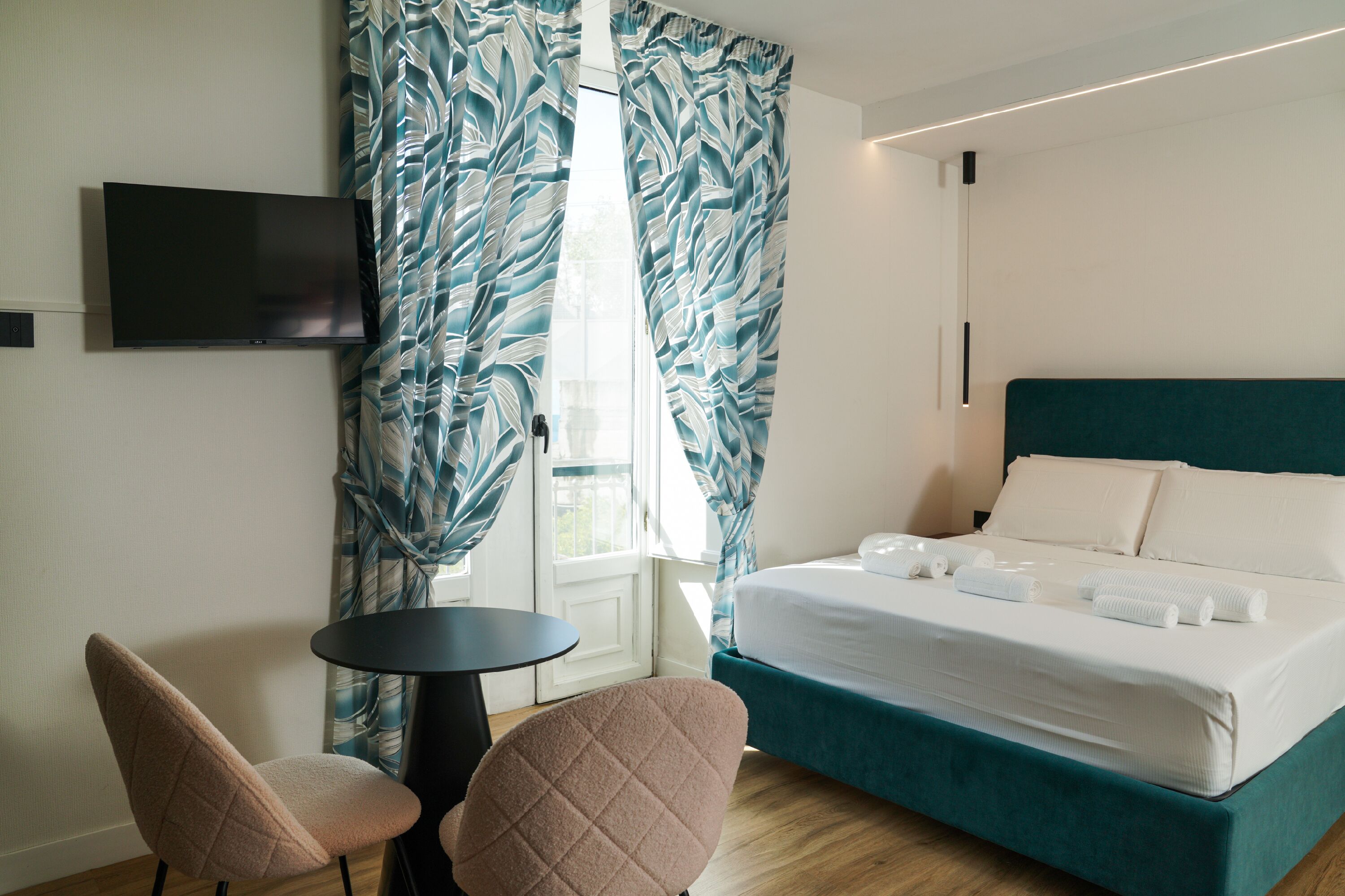 Quarto de hotel com cama, televisão fixada na parede, mesinha redonda, duas cadeiras e janela com cortinas.