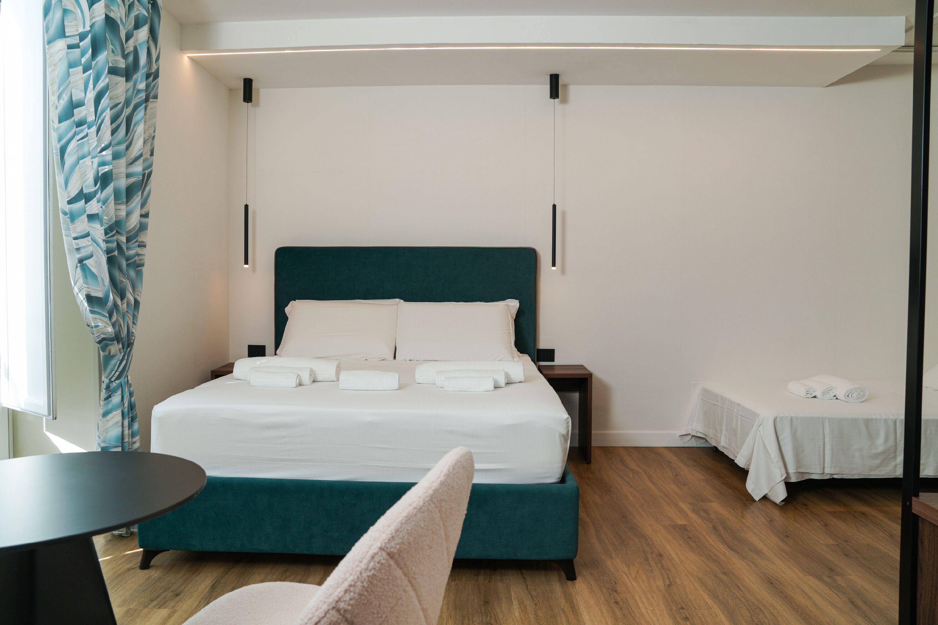 A hotel room with a bed, a small table, a chair, and a bedside table with a lamp.