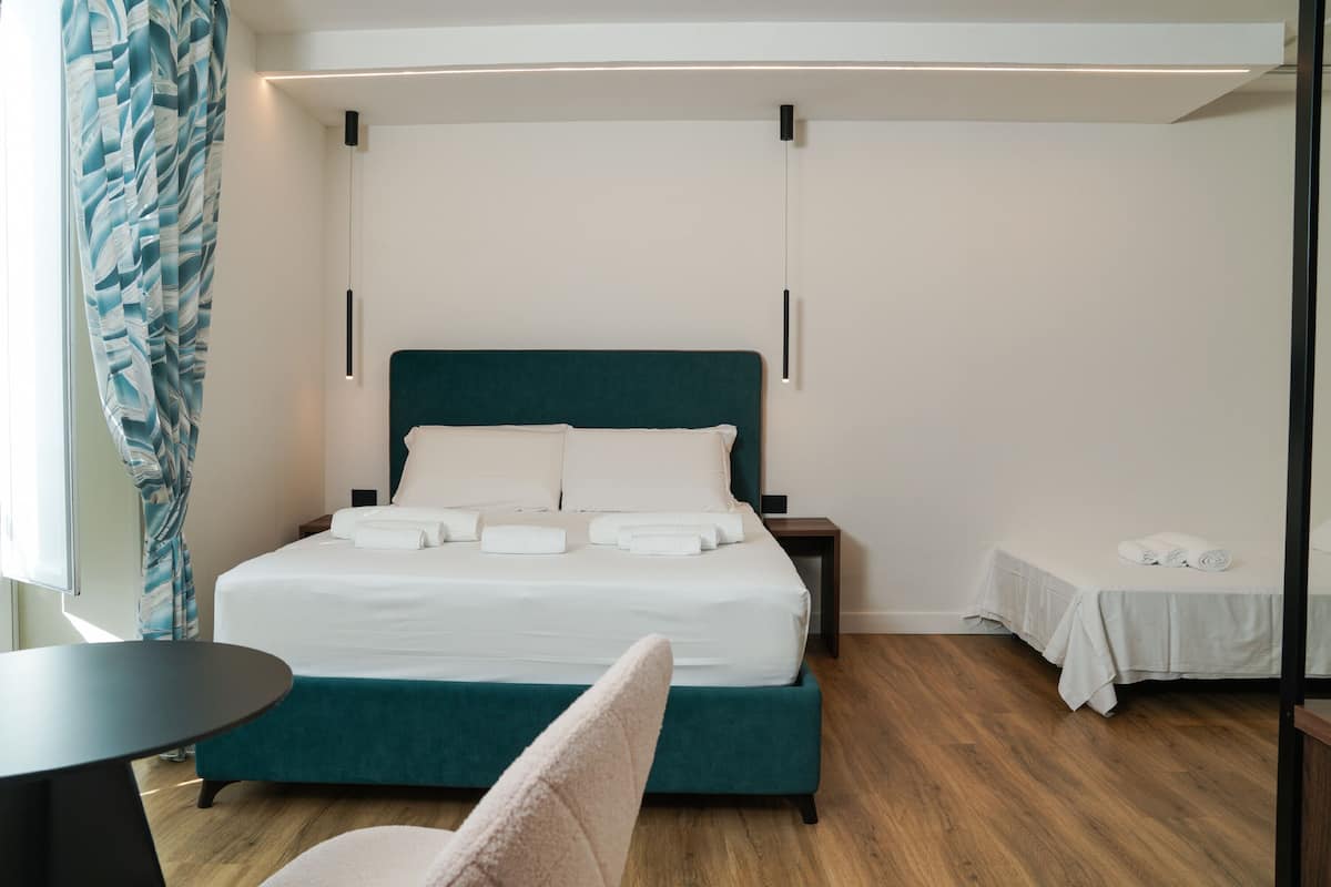 A hotel room with a bed, a small table, a chair, and a bedside table with a lamp.