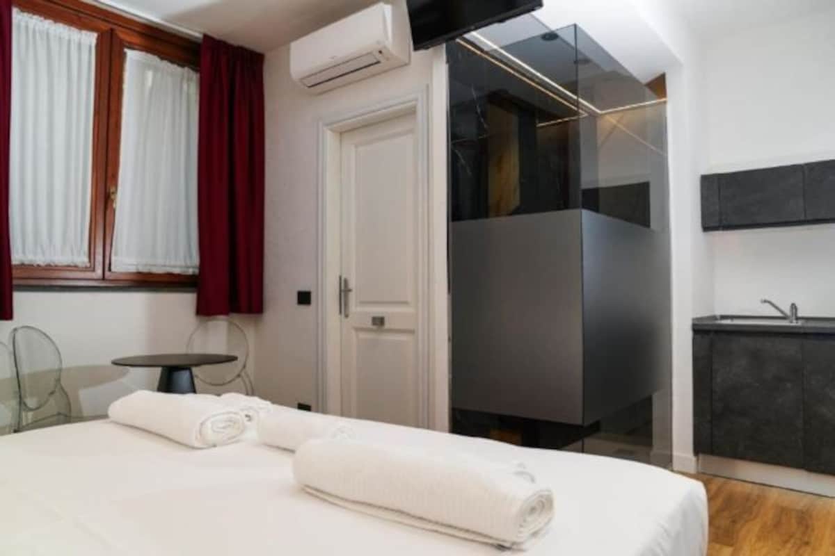 Una habitación de hotel moderna con cama, una mesita pequeña, cabina de ducha de vidrio y baño en tonos oscuros.