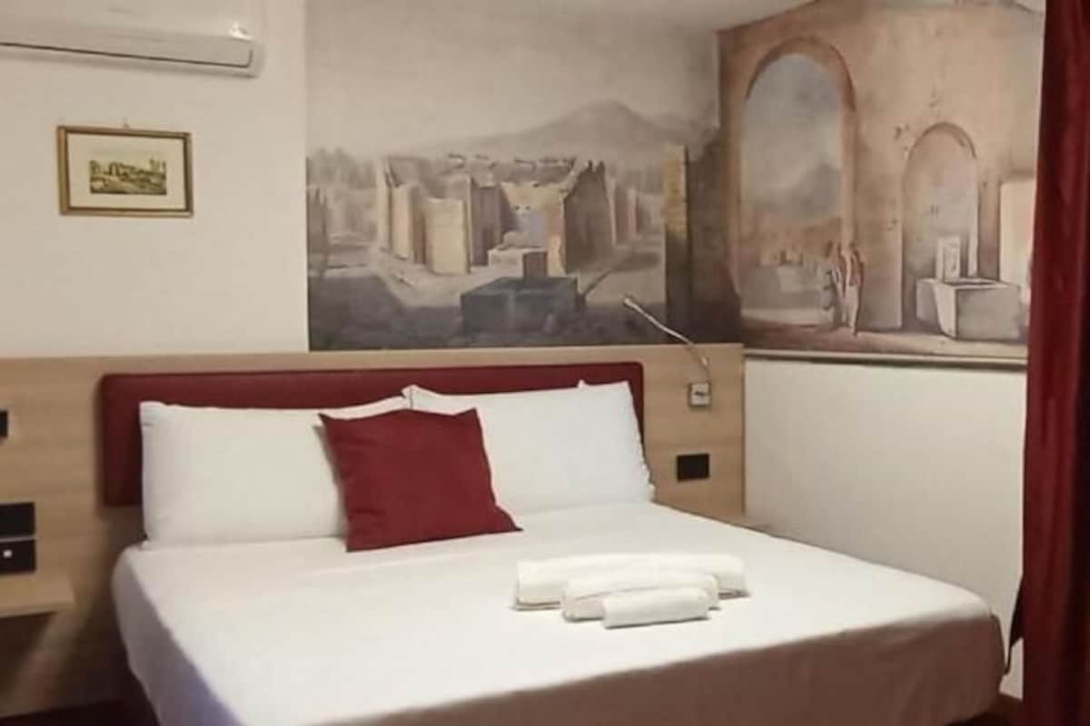 Habitación de hotel con una cama grande, cabecera roja y un mural en la pared que representa una escena histórica.