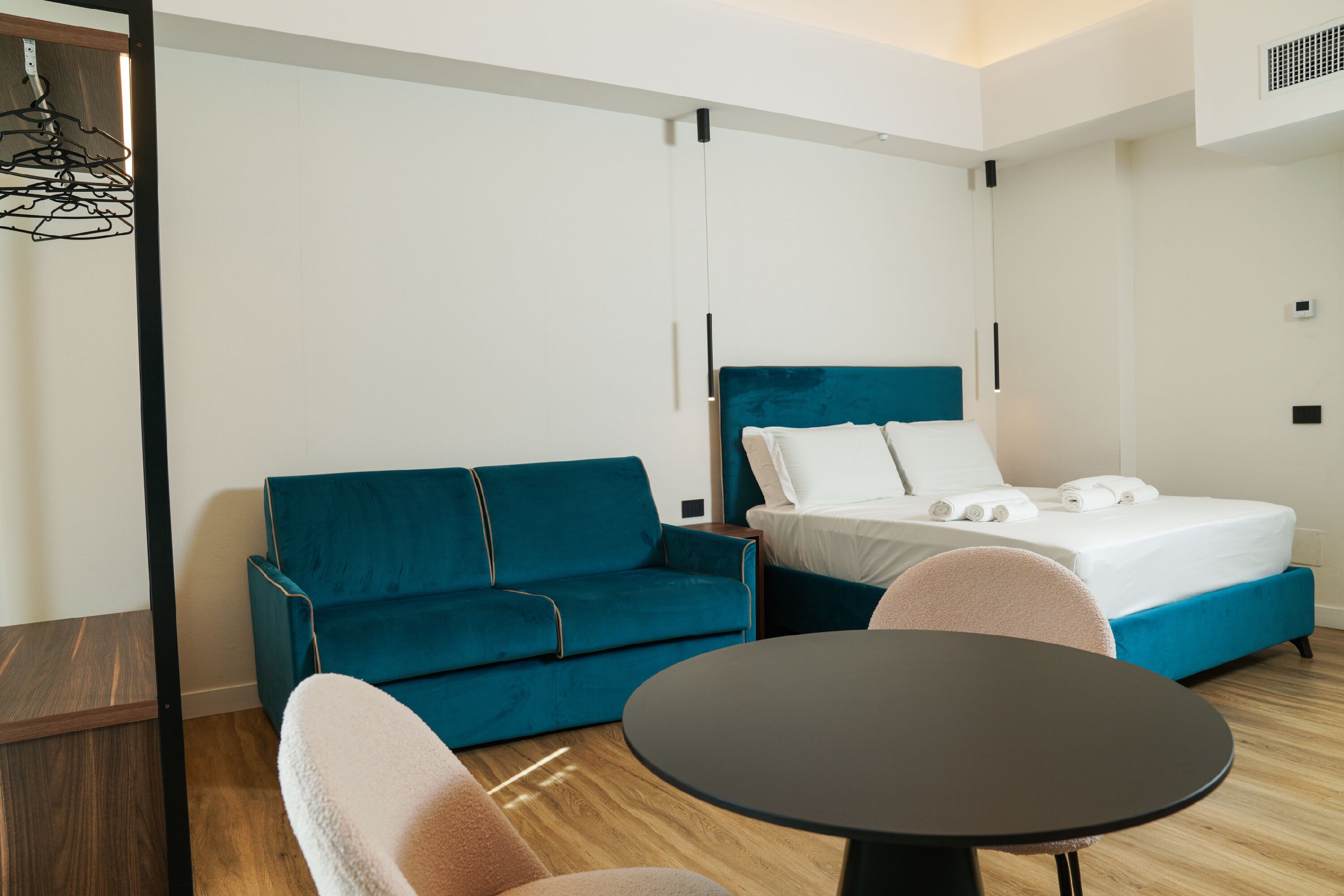 A hotel room with a teal sofa, a bed with white linens, a round black table, and a wooden floor.