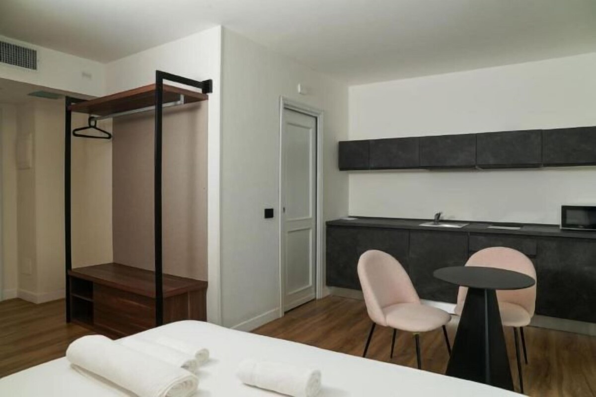 A modern hotel room with a bed, a small table, two chairs, a wardrobe, and a kitchenette.