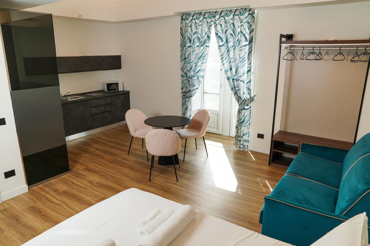 A modern room with a bed, a dining table with chairs, a sofa, a kitchen area, and a window with curtains.