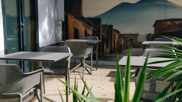 An outdoor seating area with tables and chairs, a view of a mountain, and a building with a glass door. - Hotel Il Tesoro Pompeiano (Pompei)