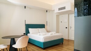 A modern hotel room with a teal bed, white bedding, a round black table, and a single white chair. - Hotel Il Tesoro Pompeiano (Pompei)