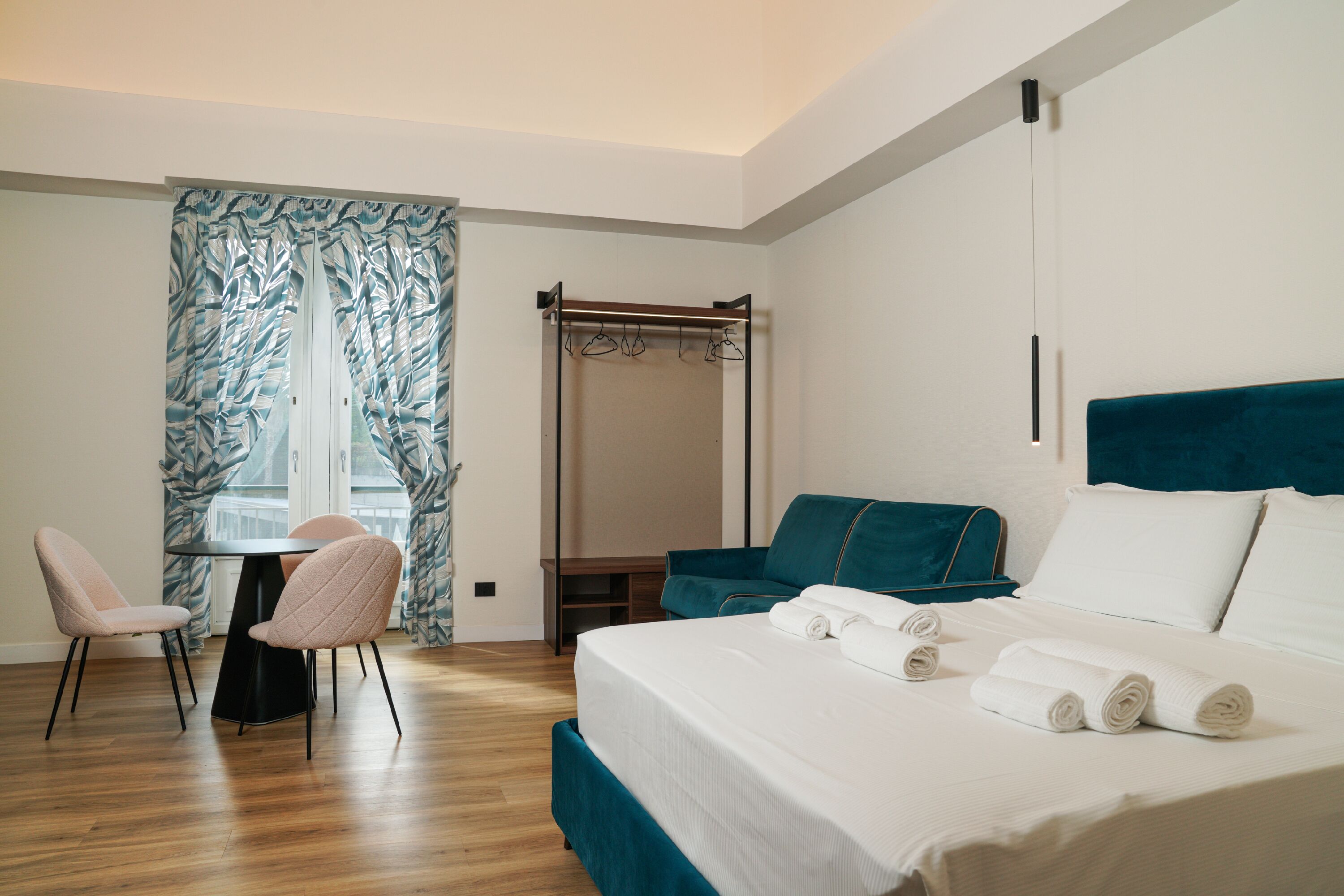 Una habitación de hotel con cama, escritorio, dos sillas, ventana con cortinas y perchero.
