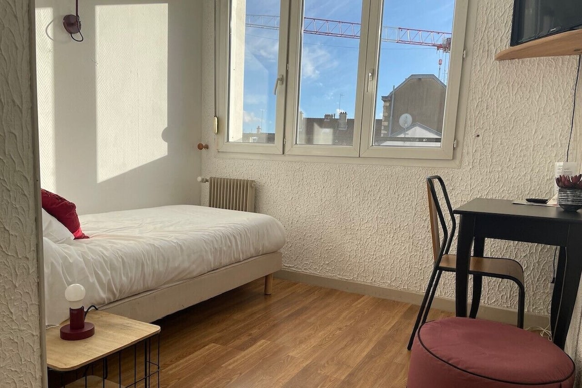 A compact room with a bed, a small table, a chair, and a window with a view of buildings.