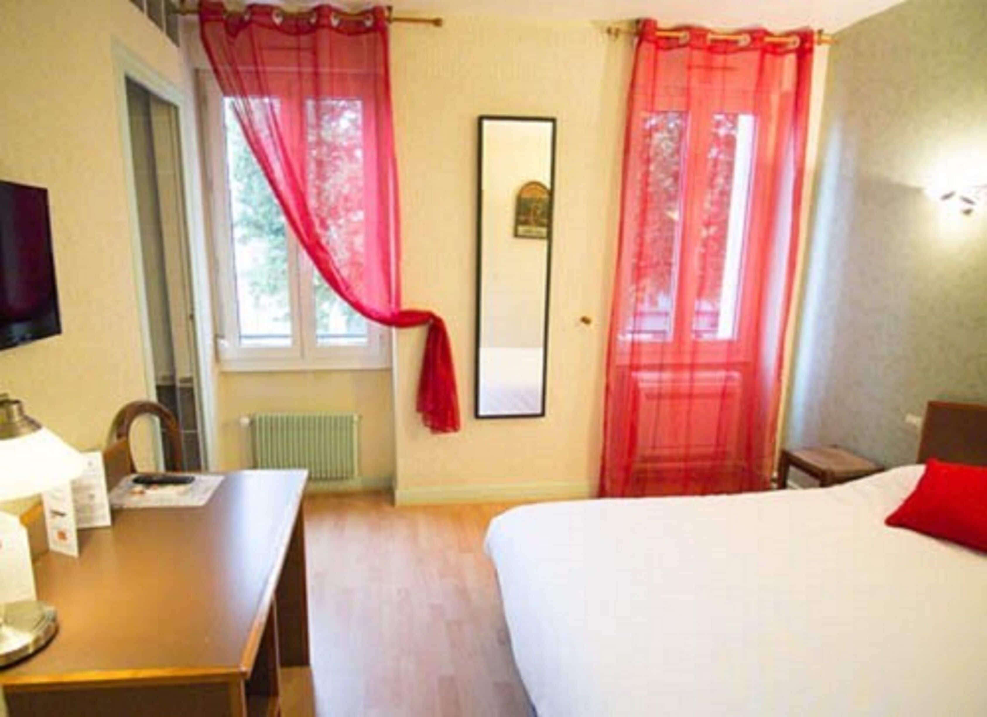 A hotel room with a bed, a desk, a chair, a radiator, and a window with red curtains.