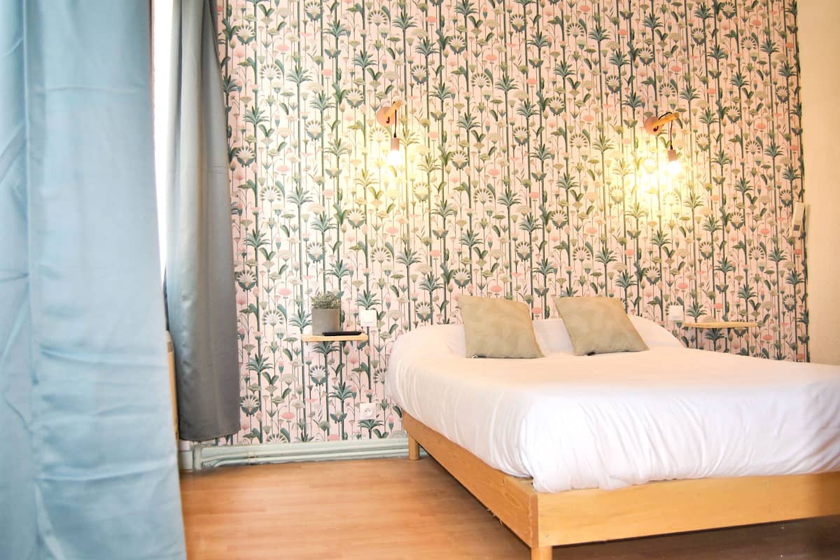 A bedroom with a floral wallpaper, a wooden bed, two bedside tables with lamps, and a window with curtains.