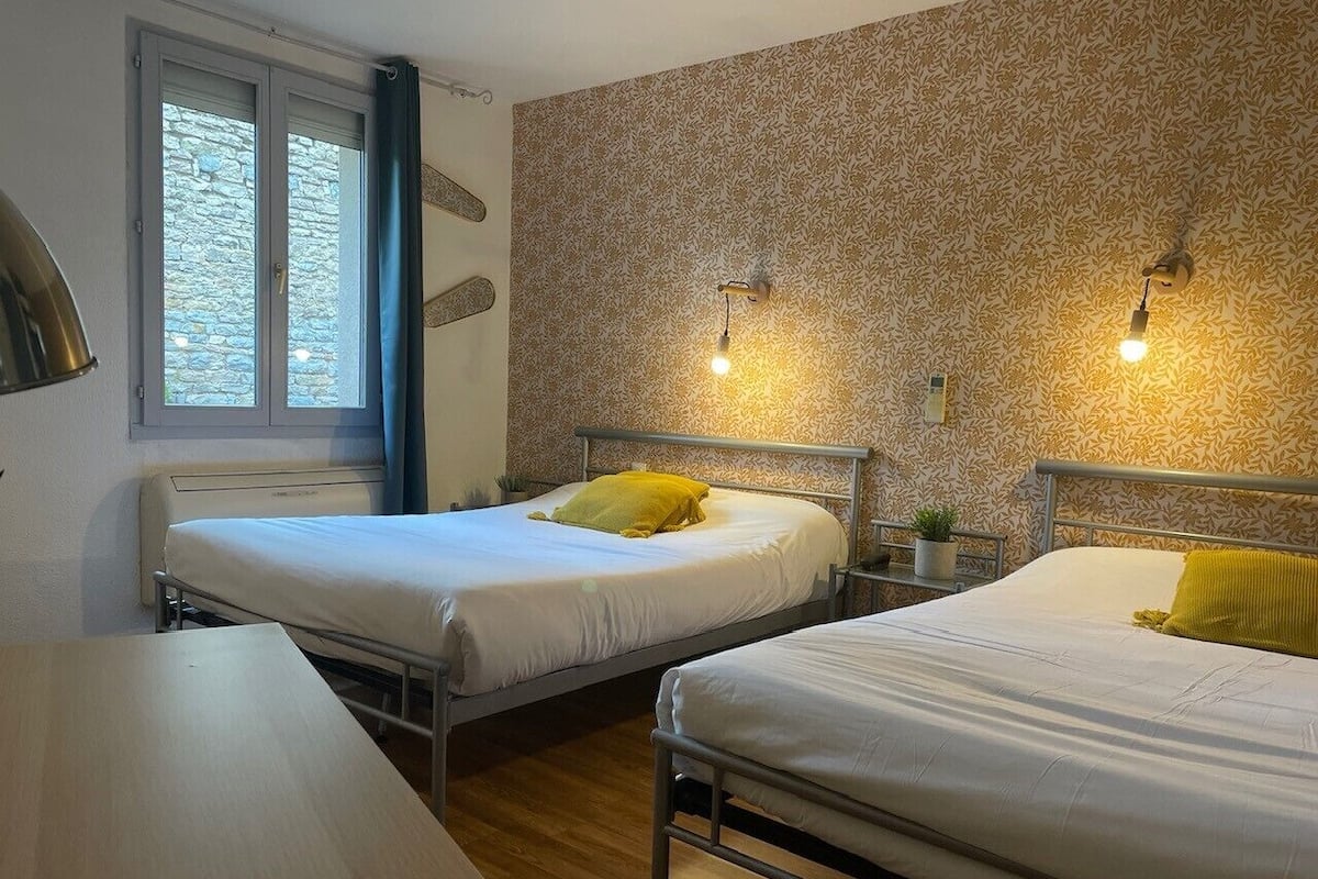 A room with two single beds, a window with curtains, a wall-mounted lamp, and a small potted plant.
