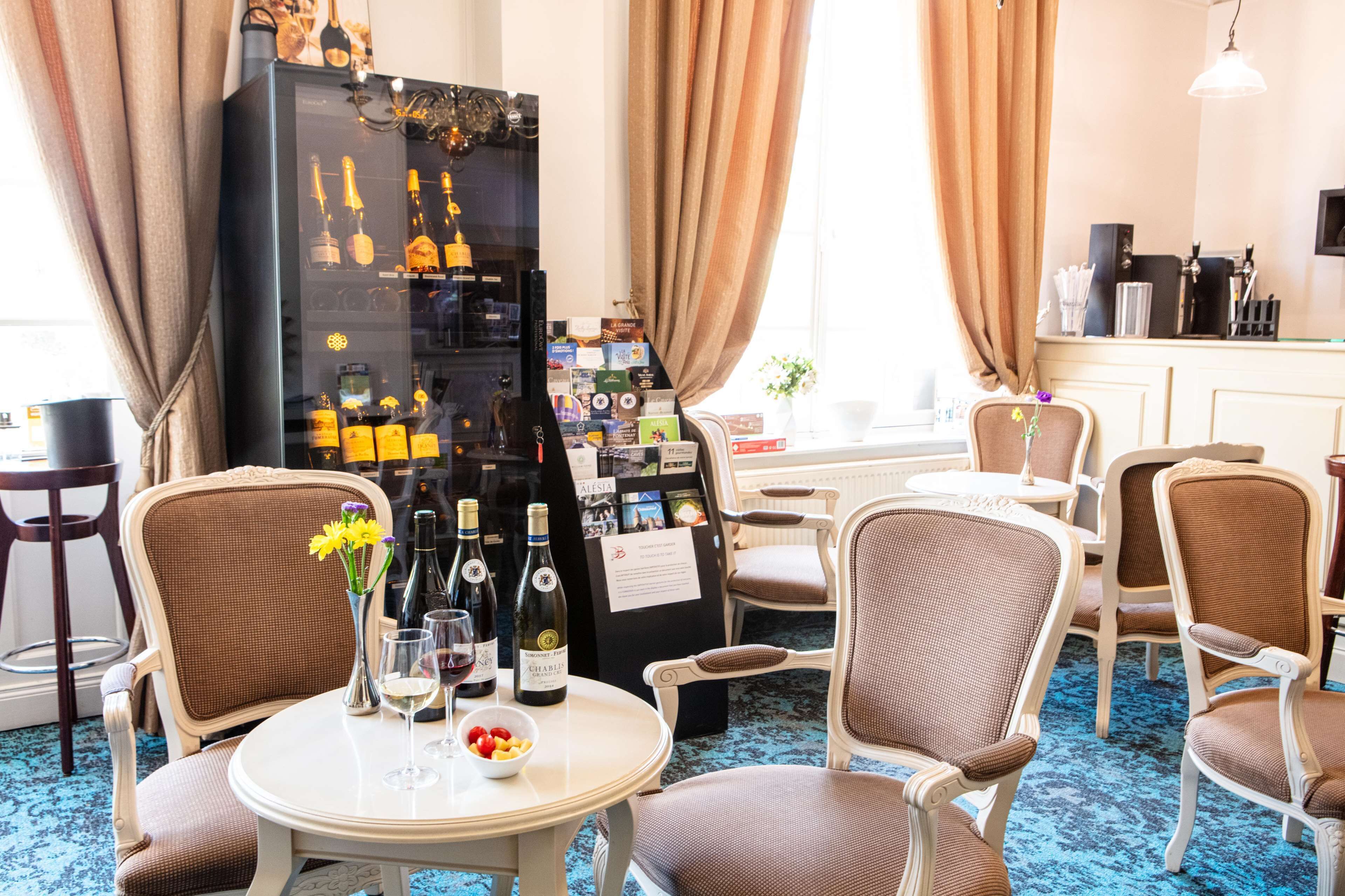 Habitación de hotel con zona de comedor que incluye una mesa redonda, varias sillas, un refrigerador para vinos repleto de botellas y vistas de la ciudad a través de amplias ventanas.