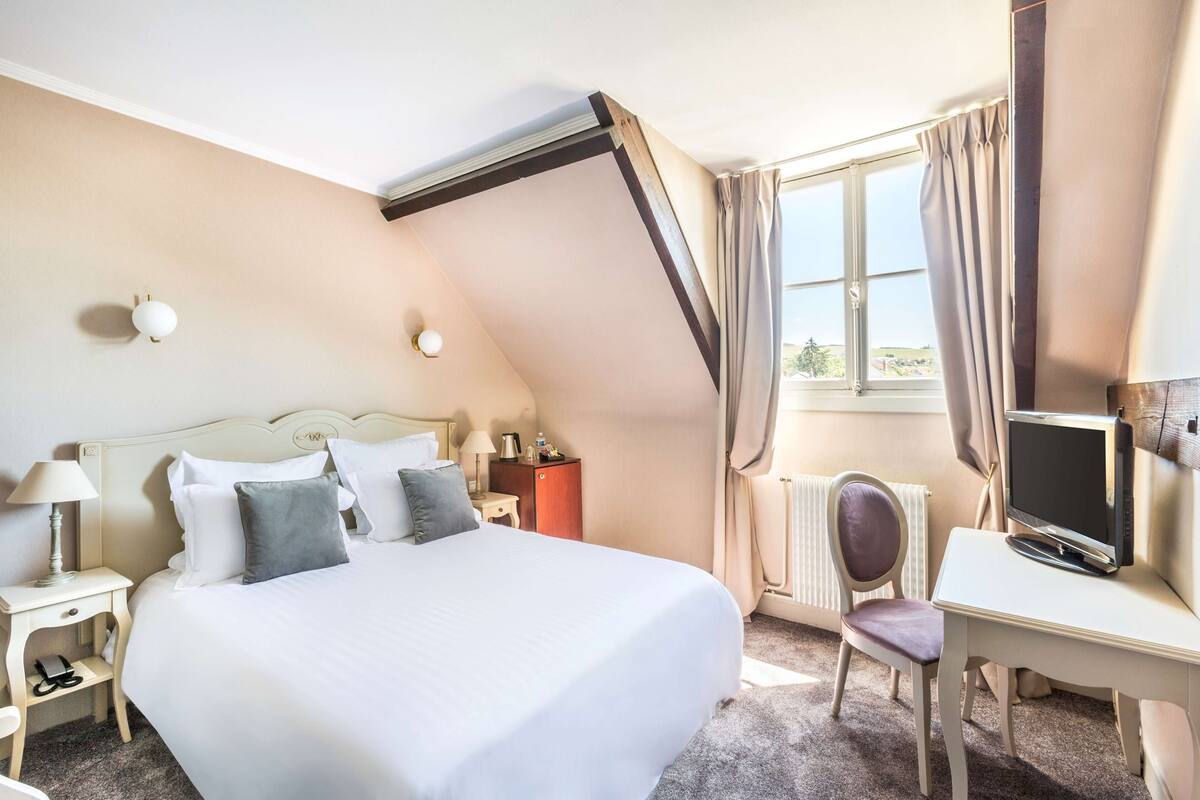 Habitación de hotel con cama, escritorio, silla, televisor, lámpara, mesita de noche y ventana con cortinas.
