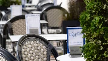 Terrassensitzplatz mit Korbstühlen und Tischen, ein Schild für 'Hôtel Maxim' und eine 'La carte du bar'-Speisekarte.