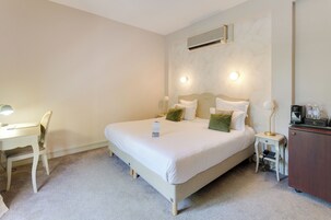 Habitación de hotel ordenada con cama, mesita de noche, lámpara y un pequeño escritorio con silla.
