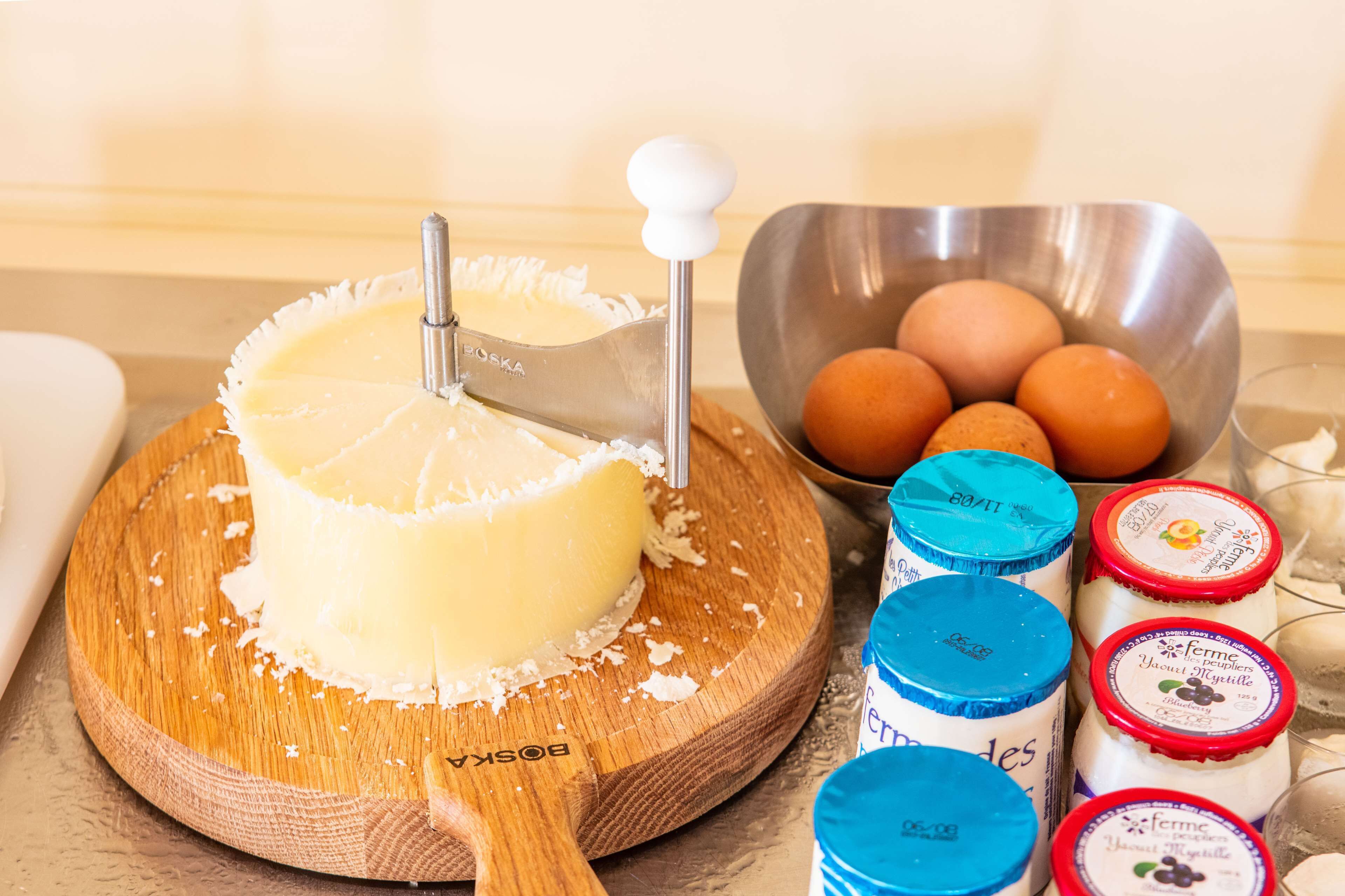 Um queijo redondo sobre uma tábua de madeira, com um fatiador de queijo, um recipiente com ovos e vários potes de iogurte.