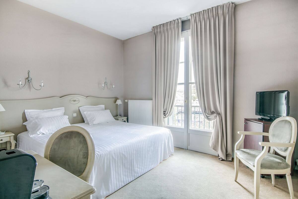 Habitación de hotel con cama, una silla, escritorio, televisión y ventana con cortinas.