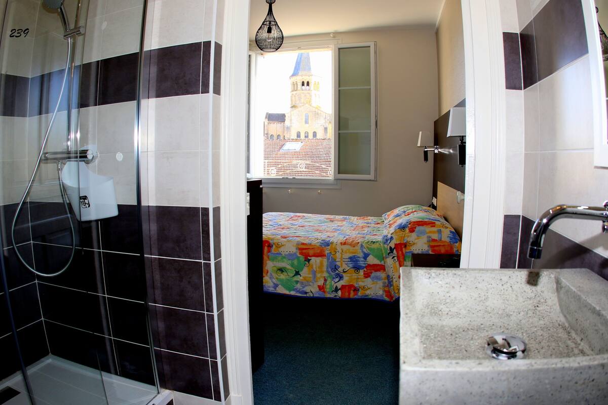 Une salle de bain avec une douche, un lavabo et un lit recouvert d’une courtepointe aux couleurs vives. Une fenêtre donne sur un immeuble doté d’une tour.