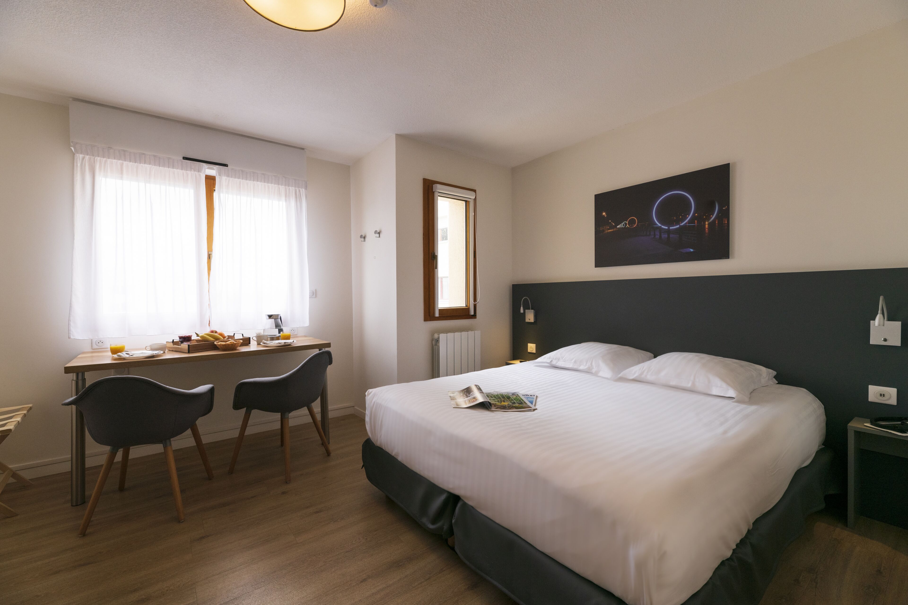 Habitación de hotel con cama, escritorio con sillas, ventana con cortinas y una obra de arte en la pared.