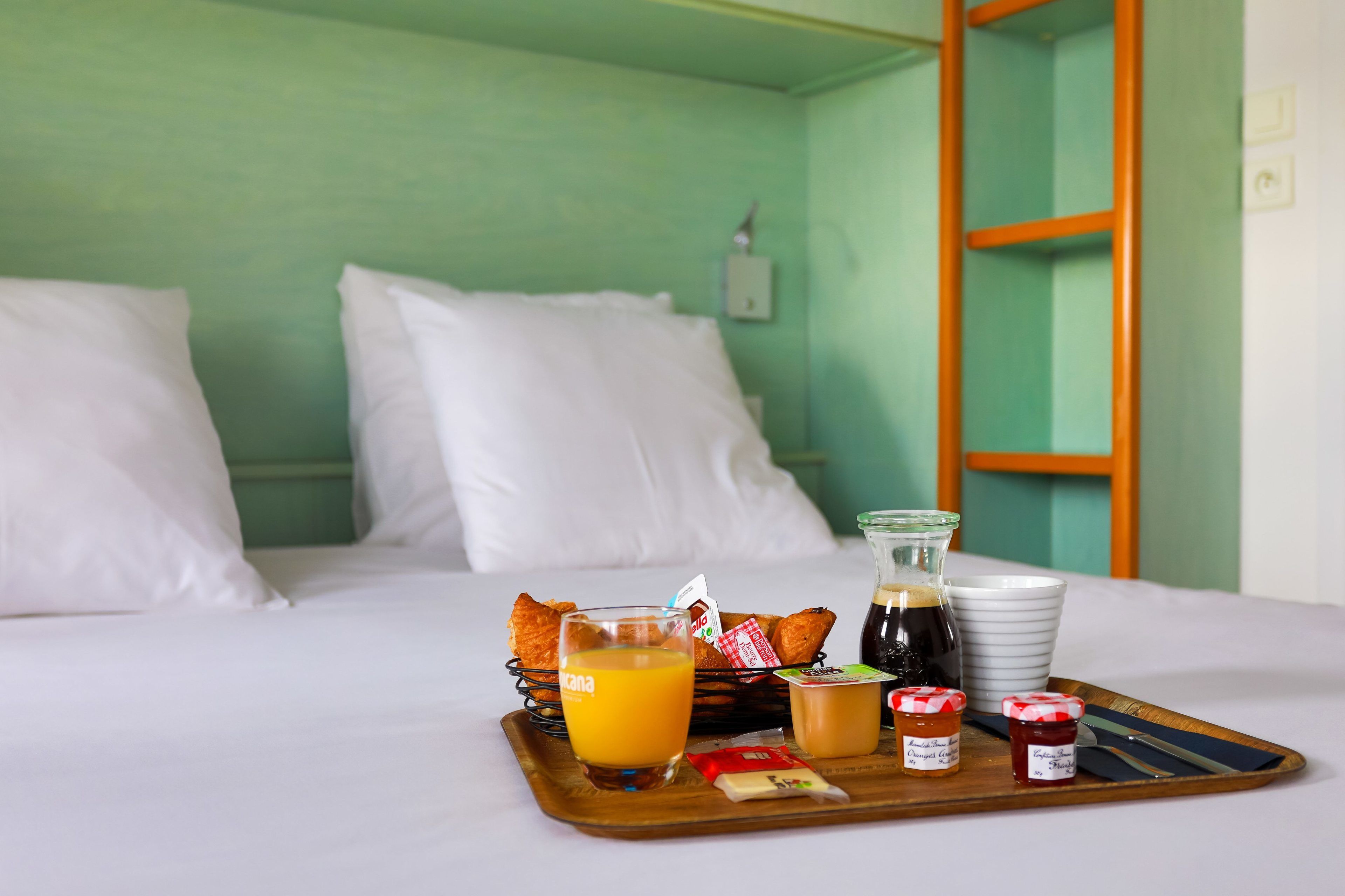 Una cama bien hecha con almohadillas blancas y una bandeja con alimentos para el desayuno, como jugo de naranja, pan, mermelada y una cafetera.